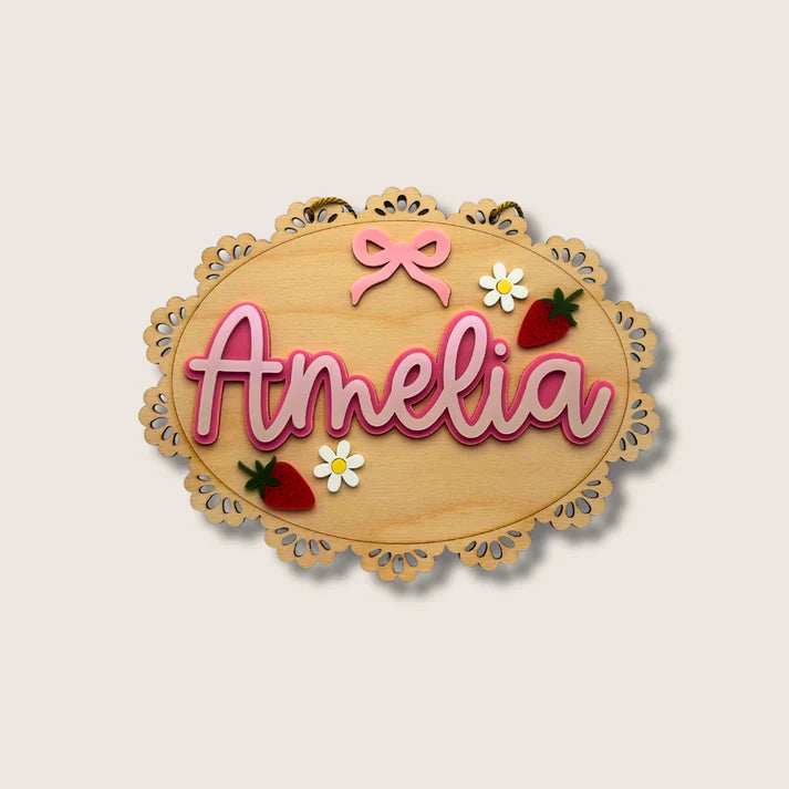 Wooden nameplate with 'Amelia' in pink, decorative elements like a bow and flowers on a beige background